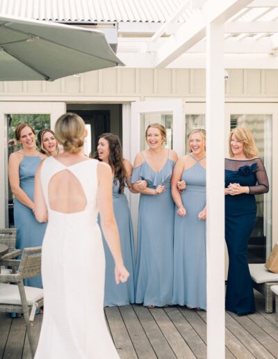 A bride walks toward a group of smiling bridesmaids in blue dresses on a wooden deck.