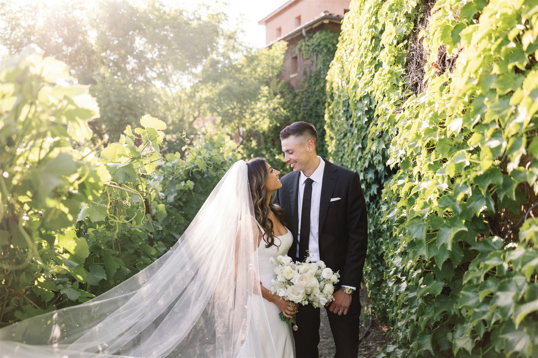 The bride in a white dress and veil, holding a bouquet, stands with the groom in a black suit and tie amidst lush greenery and ivy-covered walls, evoking an Italian-inspired wedding ambiance.