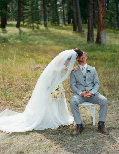 Bride in a white dress and veil kisses a groom in a gray suit seated on a chair in a forested area. She holds a bouquet of white and pastel flowers.