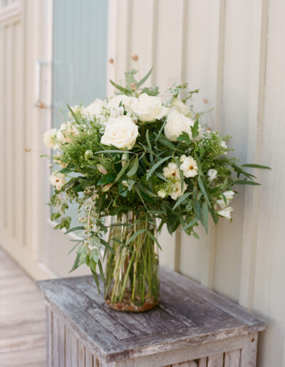 A bouquet of white roses and greenery in a clear vase is placed on a rustic wooden table against a light-colored wall.