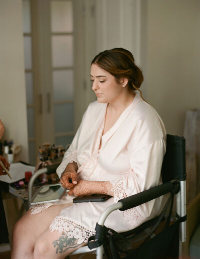 A woman in a light pink robe sits in a chair with henna on her hands. She has a floral tattoo on her thigh, and makeup items are visible nearby.