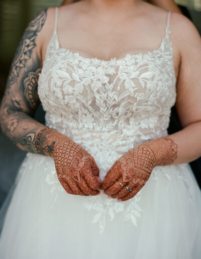 A person in a detailed white lace wedding dress displays intricate henna designs on both hands and tattoos on their arms.