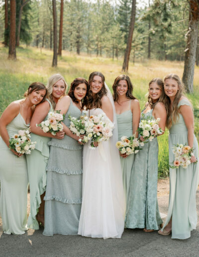 A bride and six bridesmaids in light green dresses smile and hold bouquets in a forest setting.