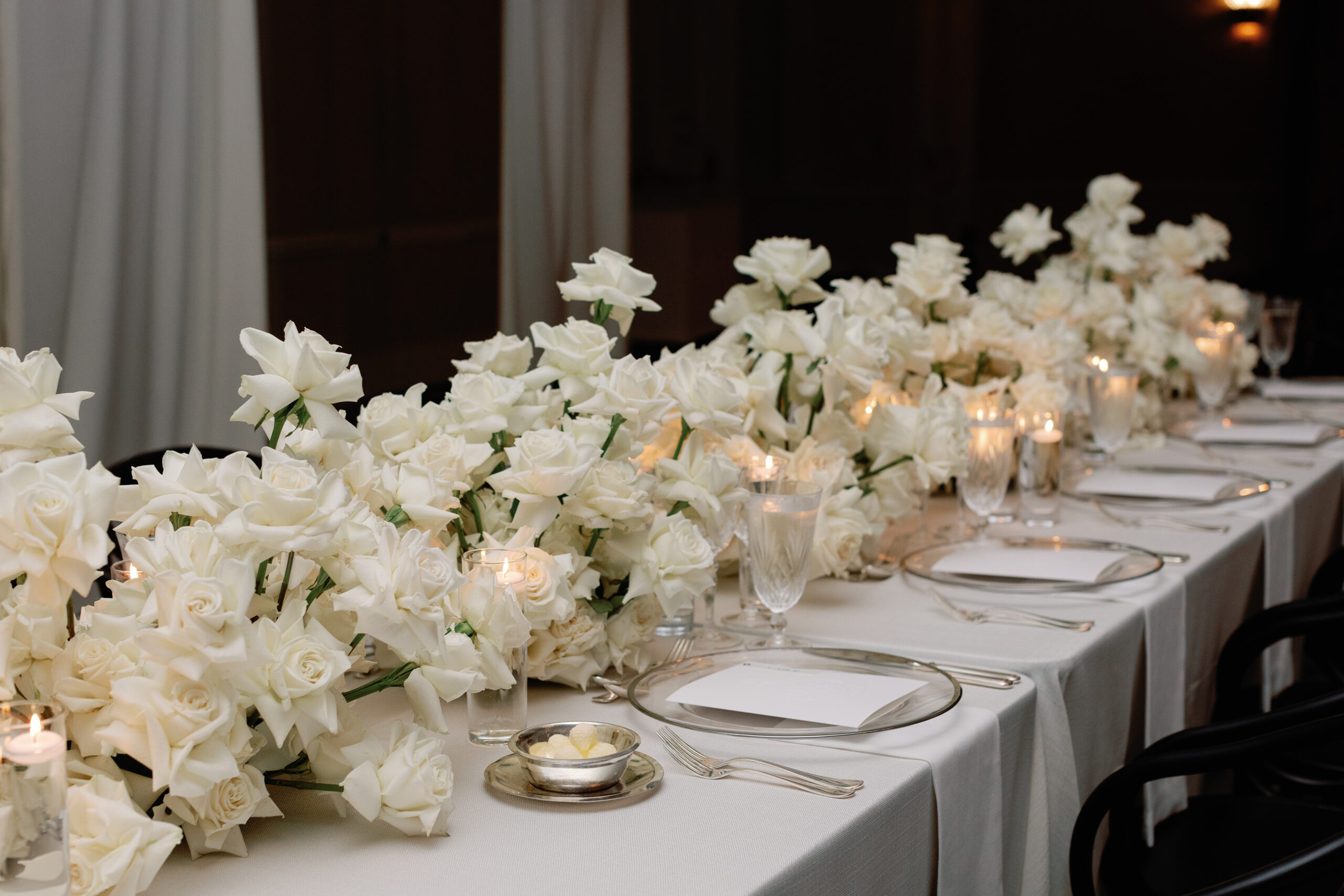 Elegant dining table setup reminiscent of a destination wedding planner's dream, with white roses, candles, plates, and glasses arranged neatly on a pristine white tablecloth.