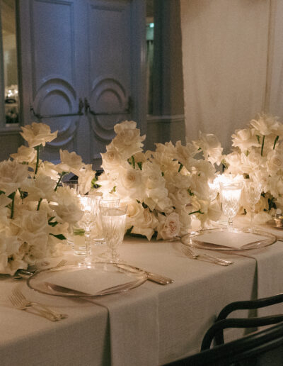 Elegant table setting with white flowers and lit candles arranged in the center. Crystal glassware, silver cutlery, and white napkins are neatly placed on a long table covered with a white cloth.