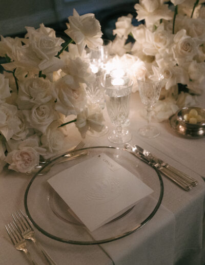 Elegant table setting with white roses, crystal glassware, silverware, and a menu card on a clear plate. Candles add soft lighting.