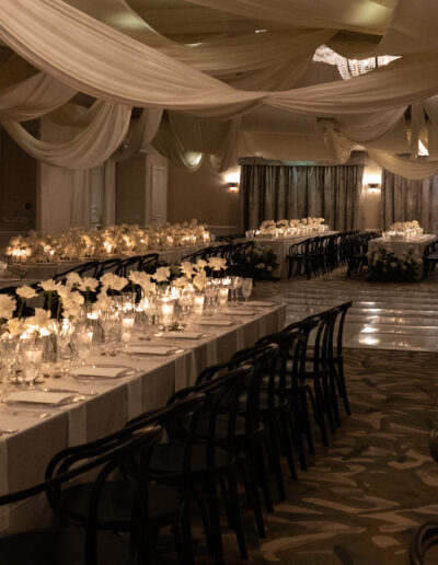 Elegant banquet hall with long tables, black chairs, white flowers, and candles. Draped ceiling adds a sophisticated touch.