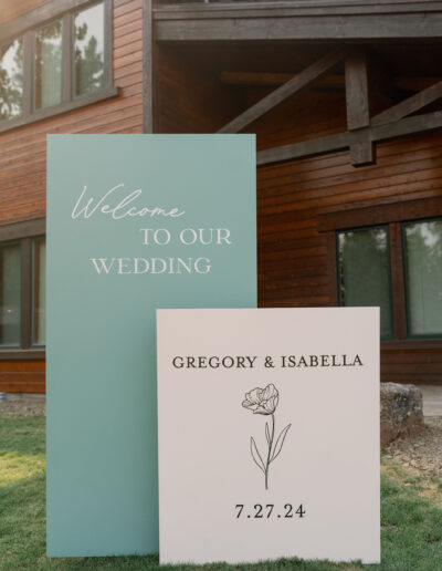 Two wedding signs: one reads "Welcome to Our Wedding," the other says "Gregory & Isabella, 7.27.24" with a floral illustration, set against a wooden building backdrop.