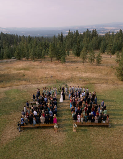 Aerial view of an outdoor wedding ceremony in a field surrounded by trees. Guests are seated on each side of the aisle as the couple stands at the altar.