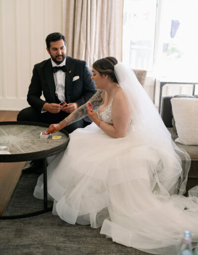 Bride and groom in wedding attire play cards at a table. The bride is in a white gown with a veil, and the groom is in a black suit with a bow tie.