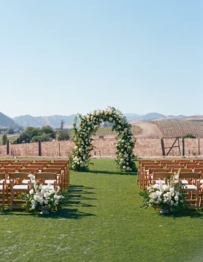 Outdoor wedding setup with wooden chairs facing a floral archway on a grassy field. Hills and vineyards are visible in the background under a clear blue sky.