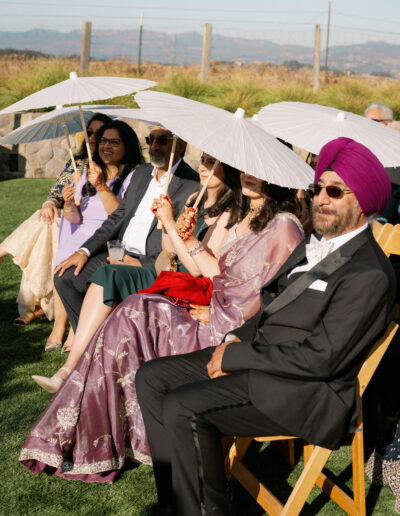 People seated outdoors, dressed formally, holding white parasols under sunny skies.