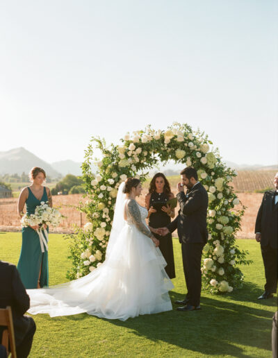 A wedding ceremony outdoors with a bride and groom standing under a floral arch. The bride is in a white dress, and the groom is in a dark suit. A bridesmaid and an officiant are present.