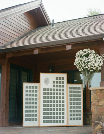 A wooden building entrance with three large seating charts displayed, featuring a logo at the top. A hanging basket with white flowers is on the right.