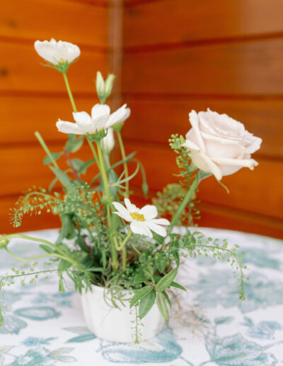 A small floral arrangement with white roses and daisies in a white vase, placed on a patterned tablecloth against a wooden wall background.