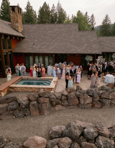 A group of people gather on a patio near a small pool outside a large wooden building surrounded by trees.