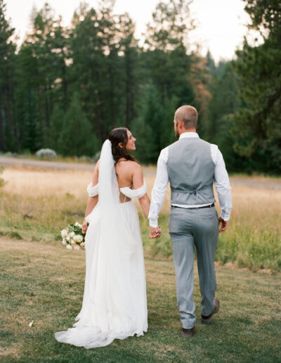 Bride and groom holding hands and walking in a grassy field, surrounded by trees.