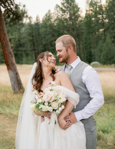 A bride and groom stand close, smiling at each other. The bride holds a bouquet of white and pink flowers. They are outdoors with trees in the background.