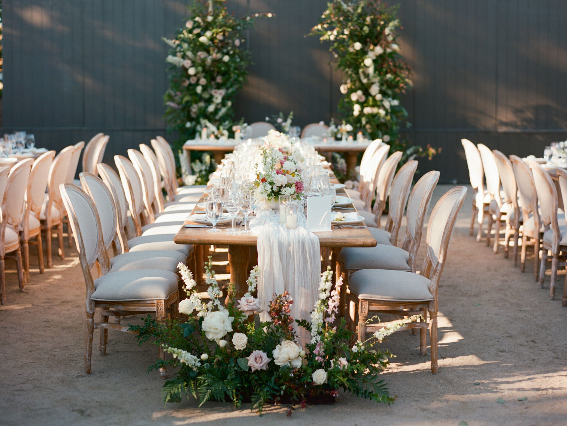 Elegant outdoor wedding reception setup with a long table, surrounded by white cushioned chairs, floral centerpieces, and greenery against a wooden backdrop.