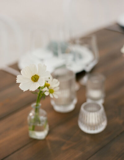 A small bouquet of white flowers in a glass vase sits on a wooden table surrounded by several empty glass cups.