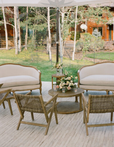 Outdoor seating area with wooden chairs and white-cushioned sofas around a round table. A flower arrangement is on the table, and trees and greenery are in the background.