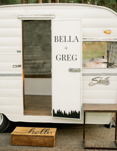 White vintage trailer with an open door displaying "Bella + Greg." A "hello" mat lies at the entrance with a step beneath it.