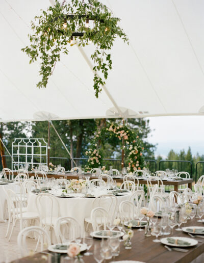 Elegant outdoor wedding reception under a tent, featuring round and rectangular tables with white chairs, floral centerpieces, and a hanging greenery arrangement.