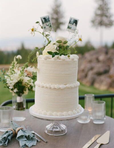 A two-tier white cake with floral decorations and photo toppers is on a table with glassware, cutlery, and floral arrangements, set outdoors with trees and rocks in the background.