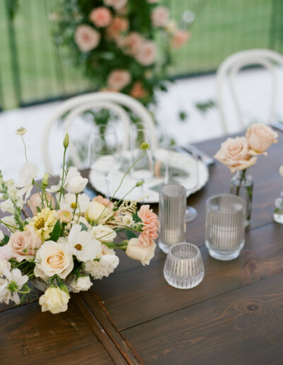 A wooden table is decorated with pastel flowers in a vase, clear glass votives, and elegant place settings. White chairs are seen in the background.