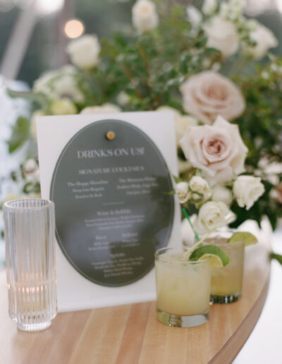 Two cocktails with lime wedges on a wooden table beside a drink menu and a vase of white roses.