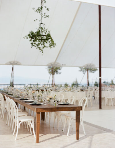 Outdoor wedding reception setup under a white tent with a long wooden table, white chairs, and hanging greenery arrangements.