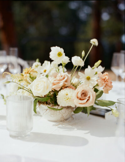 A floral centerpiece with white and peach roses, white daisies, and greenery in a bowl on a table, surrounded by glassware.