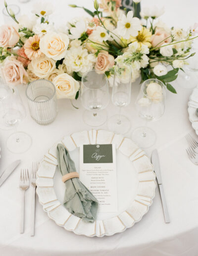 Elegant table setting with white plate, green napkin, menu card, and several wine glasses. A floral arrangement with pastel flowers is in the background.