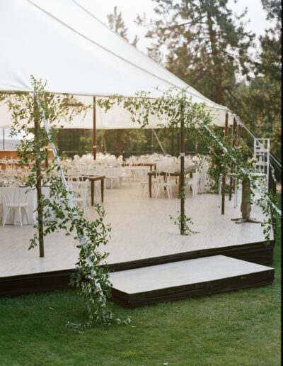 Outdoor wedding venue with a large white tent, decorated with green vines. Inside are white tables and chairs arranged neatly. Green lawn surrounds the setup under tall trees.