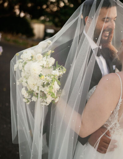 Bride holding a white flower bouquet shares a joyful moment with the groom under a veil outdoors.