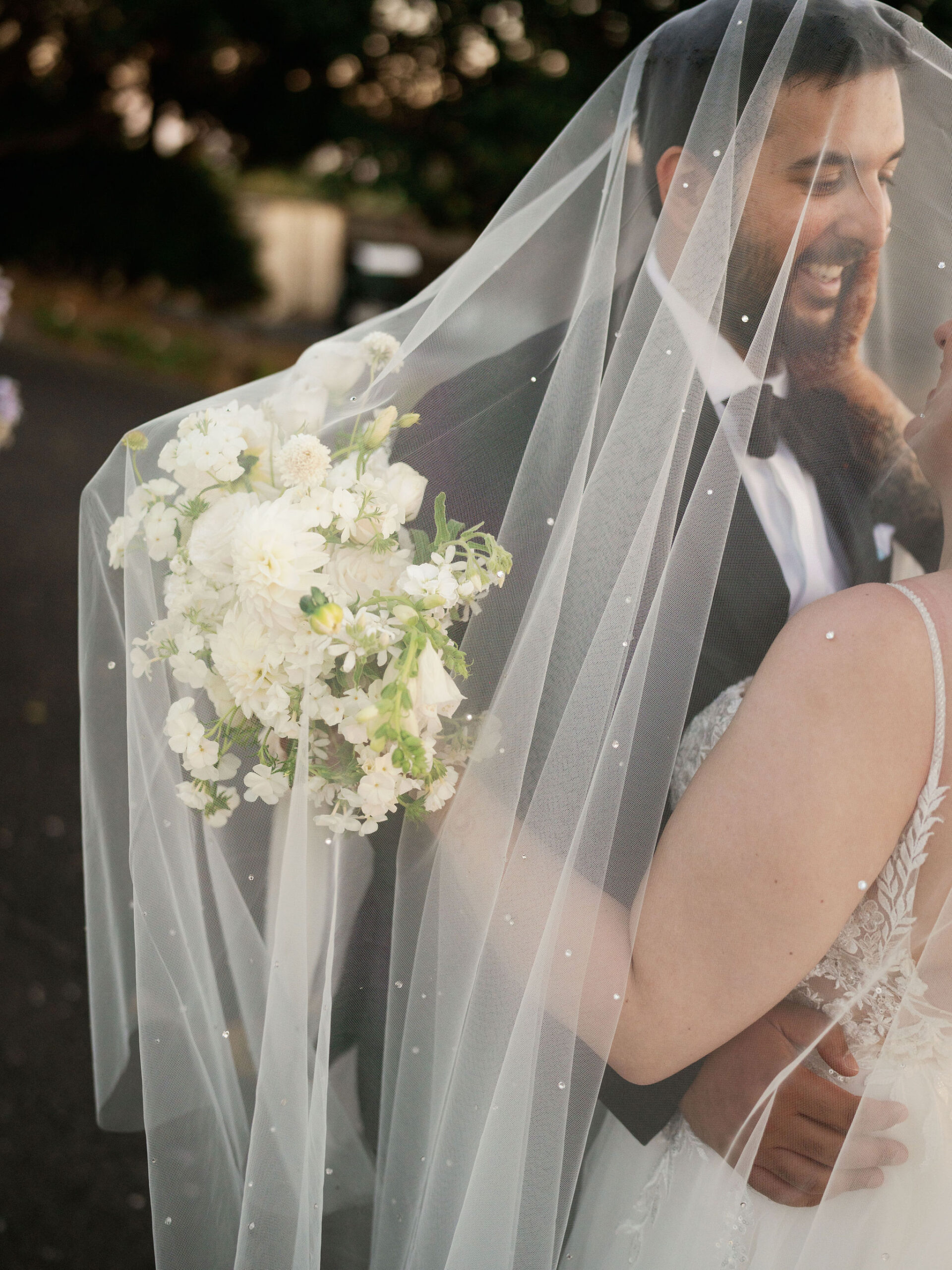 Bride holding a white flower bouquet shares a joyful moment with the groom under a veil outdoors.