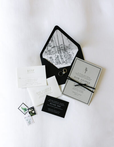 A wedding invitation suite with RSVP card, main invitation, small cards, and a black envelope with an illustrated liner. Rings and decorative fern stamps are placed nearby.