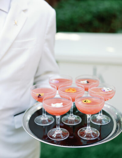 A person in a white suit holds a tray of pink cocktails garnished with colorful edible flowers.