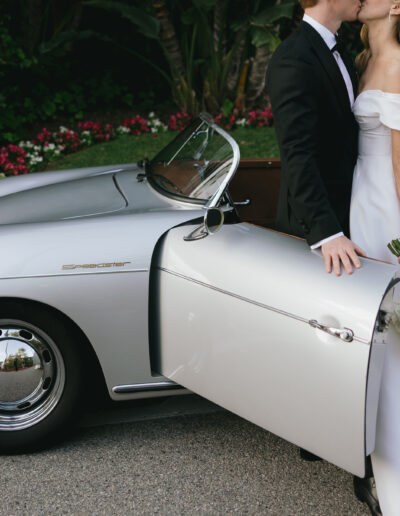 Bride and groom kissing beside a vintage silver car. Bride holds a bouquet of white roses.