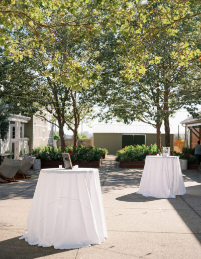 Outdoor space with two round tables draped in white cloth under tall trees. Each table holds framed photos. Buildings are visible in the background, and string lights hang above.