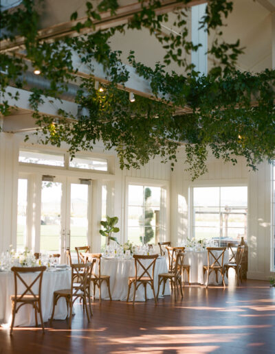 A bright room with wooden floors, white round tables set with white tablecloths, wooden chairs, and greenery hanging from the ceiling. Large windows line the walls.