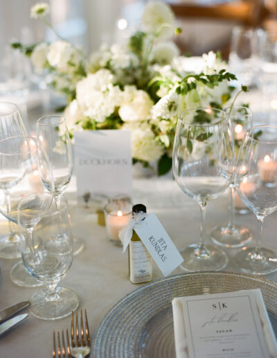 Elegant table setting with wine glasses, a floral centerpiece, and a small bottle with a tag. A menu is placed on a textured plate.