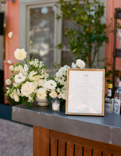 A floral arrangement next to a framed drink menu on a bar counter, with bottles and a candle nearby, set in an outdoor setting.