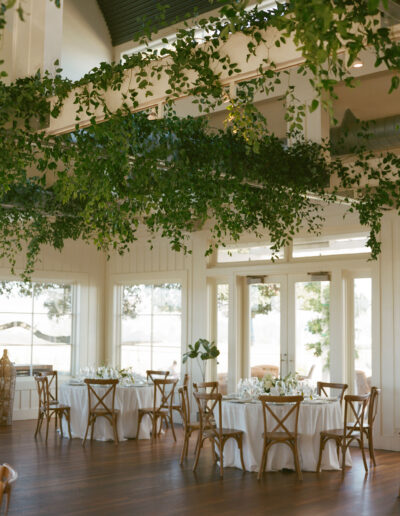 A bright, elegant dining area features round tables with white tablecloths and wooden chairs. Greenery hangs from the ceiling, and large windows offer a view outside.