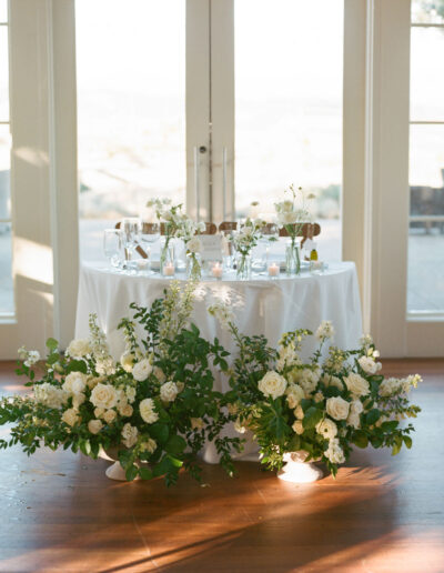 A round table decorated with white flowers and greenery is set for an event in front of large glass doors, with soft natural light streaming in.