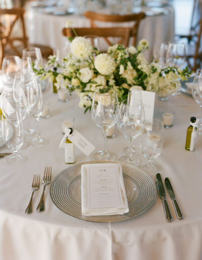 Elegant table setting with white flowers, a menu, glassware, cutlery, and name cards on a white tablecloth.