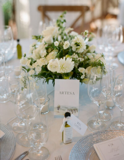 Wedding table setting with a floral centerpiece, a card labeled "Artesa," glassware, and a place card for Will Taylor on a small olive oil bottle.