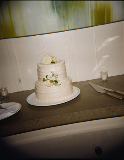 Two-tier white cake with flower decorations on a table, surrounded by a knife, fork, candles, and a glass.