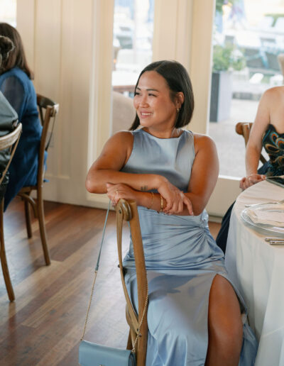 A woman in a light blue dress sits on a chair, smiling in a brightly lit room with tables set for dining.