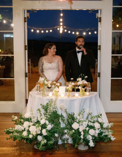 A couple stands behind a decorated table with flowers, giving a speech at their wedding reception.